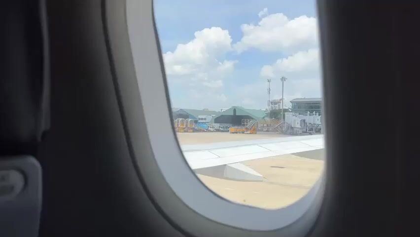 The video shows a view from an airplane window looking out at an airport tarmac. The window frame is dark gray, and the oval-shaped window reveals a bright, sunny day with white clouds in a blue sky. Outside, an airplane wing extends across the lower portion of the view. In the background, there are airport buildings, jet bridges, and other airport vehicles. The overall impression is one of a typical airport scene viewed from a stationary aircraft, likely before takeoff or after landing.