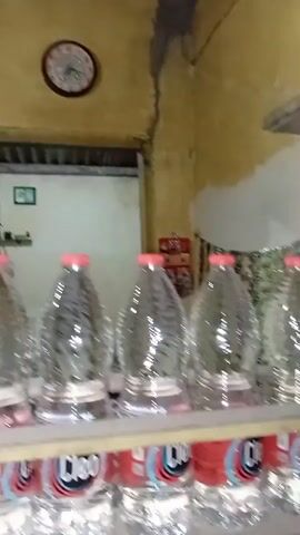 The video pans across a small, cluttered room. In the foreground, a wooden shelf holds several large, clear plastic bottles of water. The bottles have red caps and some have visible labels, one of which reads "Cleo". The shelf itself is made of light-colored wood and appears somewhat worn. Behind the shelf, the wall is painted a pale yellow, with patches of what appears to be peeling paint or dirt. A string of small, green-and-white packets hangs from the wall, possibly snacks or small goods. To the right, a white electrical box with multiple switches is mounted on the wall. Below this, a dark, rectangular box with green and red accents sits on the floor. The floor itself is a smooth, reddish-brown surface. The lighting is dim and appears to be coming from an unseen source. The overall impression is of a small store or storage area.