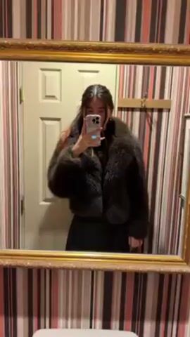 A young woman is standing in a bathroom taking a selfie in a mirror. She has long, dark hair, partially pulled back, and is wearing a dark, fluffy jacket and a dark skirt. She is holding a cell phone in her right hand, partially obscuring her face. The mirror has an ornate gold frame. The bathroom wall is covered in vertical striped wallpaper in shades of brown, red, and white. There is a white door behind her, and a rack with hooks to the right of the door. Another framed picture hangs on the wall to the right.