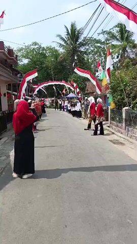 The video shows a street decorated with red and white Indonesian flags and banners, forming arches over the road. Several people are gathered, some in uniforms, others in casual attire. A group of individuals in the foreground, facing away from the camera, appear to be participating in a performance or ceremony. They are wearing red headscarves and black clothing, with some individuals in the background also in red and white outfits. The atmosphere seems festive, likely a celebration given the decorations. The setting appears to be a residential area with houses visible on the sides of the street. The sky is clear and the day is bright.