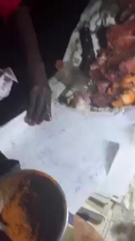 The video shows a person preparing and selling meat at a roadside stand at night.  The camera is positioned above the stand, focusing on the preparation area.  The person, who appears to have dark skin, is wearing a red shirt and using their bare hands to wrap pieces of meat in paper.  On the stand is a large pile of cooked meat, some onions, a water bottle, and skewers of meat.  A bowl of reddish-brown spice mix and a small bag are also visible on the stand.  The stand is covered with newspaper and notebook paper which is being used to wrap the meat. A ring light is illuminating the area. The environment is dark, suggesting it is nighttime.