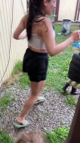 A woman and a child are in a backyard during a rainstorm. The woman is wearing a white crop top and black shorts. She is jumping with her arms raised. The child is wearing a dark vest and pants and is also jumping. There is a blue inflatable pool and a wooden ladder in the background. The ground is wet and covered in gravel and grass. A trampoline is visible behind the woman and child. The sky is overcast.