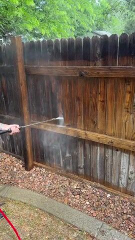 A person's left hand is visible, holding a pressure washer wand pointed at a wooden fence. The wand is spraying water onto the fence, creating a mist. The fence appears to be made of vertical wooden planks and is weathered. There is a wooden post on the left side of the fence. The ground in front of the fence is covered in small, reddish-brown rocks and some green grass. A red hose is visible in the lower left corner. Green leaves from trees are visible above the fence.