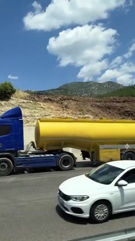 The video shows a view from a vehicle moving along a highway. In the foreground is a grey asphalt road. Behind the road is a concrete retaining wall, which appears to be a new construction. The wall is tall and stretches across the frame. Beyond the wall, the terrain rises steeply, with exposed soil and rocks. Further up the slope, a dense forest of green trees covers the hillside. Above the trees, a bright blue sky is dotted with fluffy white clouds. In the second frame, a small silver car pulling a grey trailer is visible on the road. The trailer has a metal frame and two wheels. The rest of the video continues to show the same scenery as the vehicle moves forward, with the retaining wall and forested hillside remaining visible.