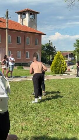 On a sunny day with a few clouds, two shirtless men in dark pants are wrestling on a grassy lawn. A building with a tower is in the background. Several people are watching the wrestling match. One man in a blue shirt is standing near the wrestlers and gesturing with his arms, likely acting as a referee. Some spectators are standing on the left side of the frame, while another man in black clothing stands off to the right.
