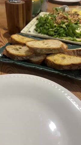 A close-up shot shows slices of toasted bread on a dark green platter. In the background, a salad with greens, walnuts, and sliced apples is on a rectangular white platter. To the right of the salad is a small white bowl with pats of butter. A blue pot sits on a straw placemat, with a ladle resting on the edge of the pot. A white plate is on the left side of the table. The table is made of wood. A woman in a patterned dress is partially visible in the background.
