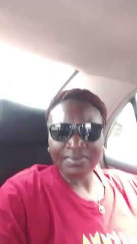 A woman with braided hair wearing sunglasses and a red "Hakuna Matata" t-shirt is inside a car. She smiles at the camera, waves, and gives a peace sign. Music can be heard.