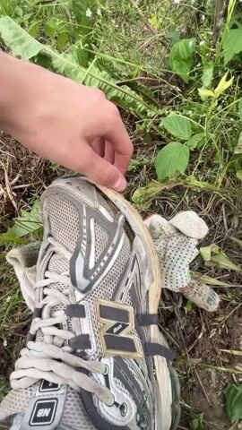 A person's hand is shown pressing down on a damaged, light-colored athletic shoe lying on the ground amidst green foliage and dry grass. A dirty, patterned glove is visible next to the shoe. The shoe appears to have a torn sole. The action is focused on the damaged area of the shoe.