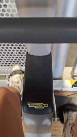 The video shows a person exercising on a Technogym stationary bike in a large, well-lit gym. The individual's legs, wearing athletic shoes, pedal back and forth as captured from a first-person perspective. The camera focuses on the Technogym logo on the bike's frame. The digital display shows workout metrics like distance, calories, and resistance level. A wider shot reveals the interior of the gym, which includes various exercise machines, weights, and other gym equipment. Several people are seen working out in the background. The gym's ceiling is exposed with visible ductwork and lighting fixtures. A perforated metal railing partially obstructs the view of the gym floor.