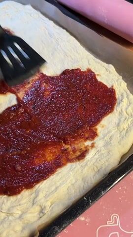 A close-up shot shows a person spreading pizza sauce onto a raw pizza crust. The crust is rectangular and has been placed on a baking sheet lined with parchment paper. A black spatula with slotted edges is being used to spread the thick, red sauce evenly across the dough. The sauce appears to have some visible herbs or spices within it. In the background, a pink rolling pin is visible, suggesting other preparations for the pizza may have occurred. The scene is brightly lit, and the focus is on the hands and the pizza-making process.