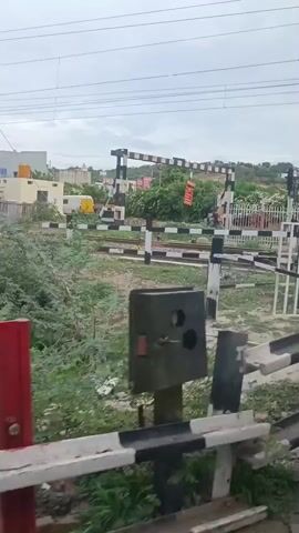 The video shows a railway crossing with barriers that appear to be closed. In the background, there are some buildings, trees, and utility poles with overhead wires. The sky is overcast. The camera is positioned to the side of the crossing, with the barriers in the foreground.
