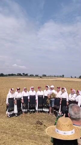 A group of approximately thirteen people, mostly women, stand in a semi-circle in a dry, grassy field under a partly cloudy sky. They are dressed in traditional folk costumes. The women are wearing long, dark skirts with decorative white blouses and headscarves. One man in the center of the group, wearing a white cap, a blue t-shirt, and denim shorts, holds a bouquet of wheat stalks. To the right, in the foreground, a person wearing a straw hat and another with a camera are visible, suggesting they are observing or documenting the scene. The background consists of a flat landscape with fields and distant trees.