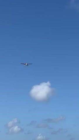 The media file captures a small seaplane in mid-flight against a clear blue sky. The plane, a twin-engine turboprop, is white with red accents on its wings and tail. It appears to be at a moderate altitude, with fluffy white clouds scattered below. One particularly large, cumulus cloud is visible directly beneath the aircraft. The bright sunlight suggests it is daytime, likely midday, and the clear sky indicates fair weather. The scene is set in an open sky, with no visible ground features. The overall mood is serene and peaceful, evoking a sense of travel or exploration over an aquatic environment, which is implied by the presence of a seaplane and given the location context of Malé, Maldives. There is no visible text in the media file.