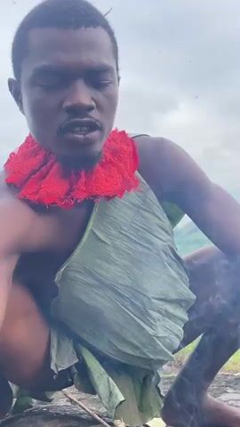 This media file features a close-up shot of a dark-skinned man in an outdoor setting. He is crouched down, appearing to be tending to a small fire in front of him. The man has a red, ruffled fabric around his neck and is wearing a garment made of large green leaves draped around his waist. His eyes are wide open, and his mouth is open as if he is blowing on the fire, creating visible smoke. The fire consists of small sticks and embers on a rocky surface, with dry grass and twigs scattered around.

The background suggests a natural environment with tall green grass and a slightly overcast sky, indicating it is daytime. The man's expression seems focused and intense, possibly due to the effort of blowing on the fire or the nature of the activity itself. The overall atmosphere is one of engagement with a primitive or traditional practice. The specific location is identified as Amaigbo, Nigeria. There is no visible text in the media file.