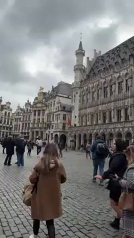 The video shows a view of the Grand Place in Brussels, Belgium, on a cloudy day. The camera pans up to show the facade of the Brussels City Hall. People are walking around in the square. The square is paved with cobblestones. Two women stand in the foreground.