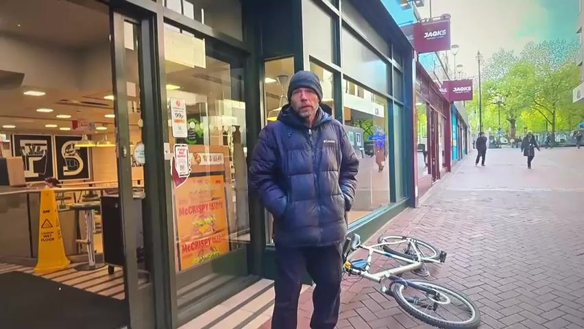 A man stands on a paved sidewalk in front of a McDonald's restaurant. He is wearing a dark blue puffer jacket, dark pants, and a dark beanie. His hands are in his jacket pockets. A bicycle lies on its side on the sidewalk behind him. The McDonald's storefront has large glass windows and a green-colored frame. Inside the restaurant, a yellow "wet floor" sign is visible. To the right of the McDonald's, there are other storefronts with signs that read "JACKS." Further down the sidewalk, other people are walking. The sky appears to be overcast.