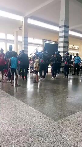 A marching band, consisting of young people in uniform, performs in a large, open indoor space with a high ceiling and natural light filtering in from windows. The floor is polished concrete. Several individuals are playing brass instruments, including trombones, and drums are visible. A group of onlookers, including children, watches the performance. There is a stage with equipment at the far end of the space.