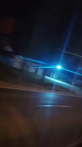 The video captures a nighttime scene from a moving vehicle. The camera is angled slightly downwards and to the right, showing a road with white lane markings. Streetlights cast bright, streaky beams of light across the frame, creating a blurred effect due to the motion. In the background, there are glimpses of trees and structures, also appearing blurred. The overall impression is of a drive through a suburban or semi-rural area at night, with the motion blurring the details of the surroundings.