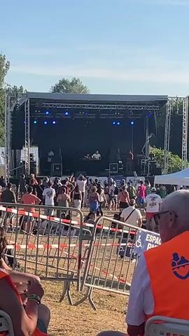 A wide shot captures an outdoor music festival from behind a crowd of attendees. In the foreground, two individuals wearing bright orange safety vests are seated, facing away from the camera. A metal barricade, strung with red and white caution tape, separates the seated individuals from the main festival area. Beyond the barricade, a large crowd of people stands and dances in front of a raised stage. The stage is equipped with a black backdrop, a sound system, and blue stage lights. Two figures are visible on the stage, one appearing to be a DJ at a console, and another standing to the right. The festival grounds are grassy, with trees visible in the background under a clear blue sky.