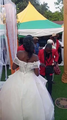 A bride in a white gown is walking onto the grass at an outdoor celebration with a yellow, green and white tent in the background. A man in a red suit and black pants is dancing with cash in his hand. He is wearing a black purse. Several men are standing around watching and recording with their phones.
