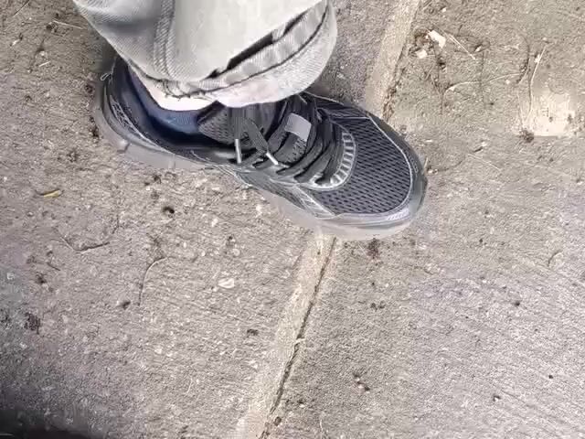 The video shows a close-up view of a person's feet wearing dark sneakers and light-colored pants. The person is walking on a concrete surface that appears to be a sidewalk or path. The camera is positioned to look down at the feet, giving a first-person perspective of the walk. Debris, such as small pebbles and dry leaves, is scattered on the concrete. The lighting suggests it is daytime.