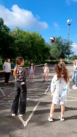 A group of children are playing volleyball on a paved court. The court has painted lines, forming a diamond shape. In the background, there are trees, a building, and a cell tower. The sky is blue with some clouds. A girl in a black and white patterned shirt is in the foreground, facing away from the camera. Another girl in a white and blue patterned tracksuit is in the center, also facing away. A girl in a light blue hoodie is to the right, facing towards the ball. A girl in a pink shirt is in the background, to the left. A girl in a purple dress is further in the background. Two other children are also visible in the background. The ball is in the air.