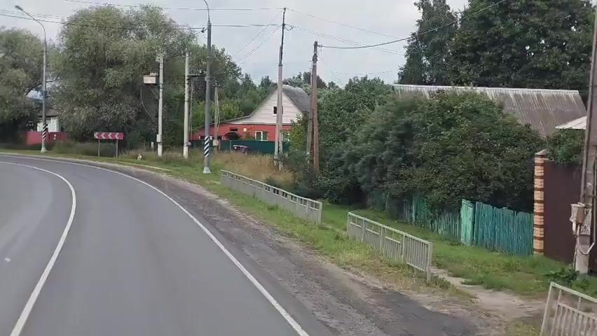 The video shows a rural road with a curve. On the left side of the road, there is a large tree and a traffic sign indicating a sharp turn. Further down the road, a red truck is driving by. On the right side of the road, there are houses and trees. A fence runs along the side of the road. The sky is overcast.