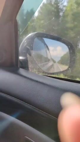 This is a first-person perspective video of a person driving a car on a sunny day. The video focuses on the side-view mirror, which shows the road ahead, the sky with white clouds, and trees lining the road. The interior of the car, including the car door panel, is also visible. The car appears to be on a paved road, and the scenery suggests a rural or suburban area. The video is brief, capturing a few seconds of the journey.