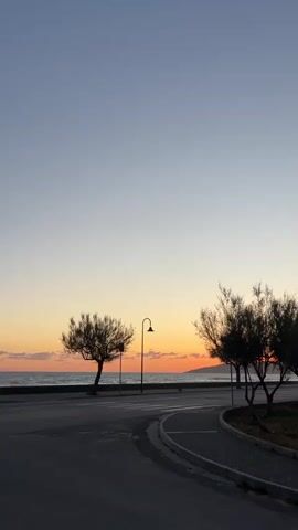 The video shows a seaside view at dusk. The sky transitions from a pale blue at the top to a gradient of orange and pink near the horizon, where the sun is setting over the water. A dark coastline is visible in the distance. The sea is calm with gentle waves. A road curves along the shoreline, and a blue car drives along it from left to right. On either side of the road are small, dark trees with sparse foliage. A vintage-style lamppost stands along the roadside. The road surface is dark, and there is a narrow strip of gravel or concrete along the edge closer to the trees. The video is still, with the only movement coming from the car.
