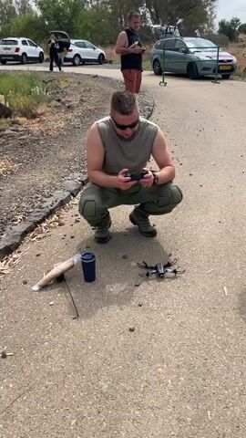 A man wearing sunglasses, a sleeveless shirt, and tactical pants is crouching on an asphalt road, holding a drone controller. In front of him, a small drone is on the ground, next to a blue cup and a long, thin object wrapped in plastic with a stick. Another man is standing a few feet behind him, also holding a controller, and walking slowly forward. In the background, several cars are parked on the side of the road, and two people are near one of the cars. The scene appears to be outdoors in a rural or semi-rural area with dry vegetation and some trees.