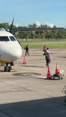 The video captures an outdoor scene at an airport tarmac in Del Carmen, Philippines. A white turboprop aircraft, possibly a passenger plane, is parked with its nose facing the viewer. The number "87" and partial text "BY: INC" are visible on its side. Several people are walking on the tarmac: two individuals with backpacks are walking away from the aircraft, while another person in a high-visibility vest with "DUTY" written on it and another person carrying a child walk towards the background. Orange traffic cones are placed near the aircraft and a small cart with more cones is also visible. In the background, there are lush green trees, including palm trees, under a bright blue sky, indicating sunny weather. The overall impression is an active airport environment during the daytime.
