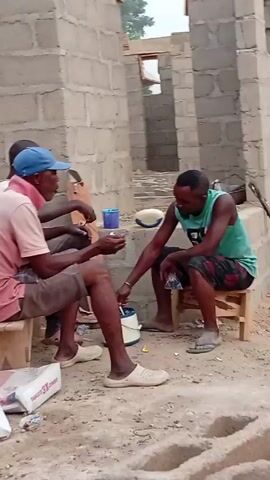 Three men are seated on small wooden stools at a construction site. The man on the left wears a pink shirt, brown shorts, a blue baseball cap, and cream-colored slide sandals. He holds a blue mug. Another man sits to his left, mostly obscured. The man on the right wears a green tank top and patterned shorts, and he's also wearing slide sandals. He reaches out to touch the wrist of the man with the pink shirt. A white bucket and a torn bag of "DANGOTE CEMENT" are on the ground. The construction site has walls made of concrete blocks.