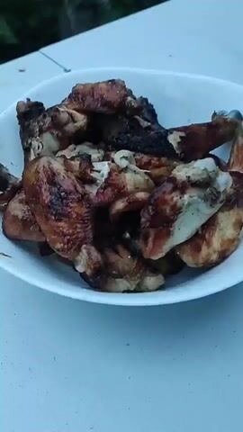 The video shows a bowl of cooked chicken wings on a white table. Next to the bowl of chicken are sliced cucumbers on a plate. In the background, there are trees and greenery. The video also includes cutlery on the table and another white plate with food on it. The lighting suggests it is daytime.
