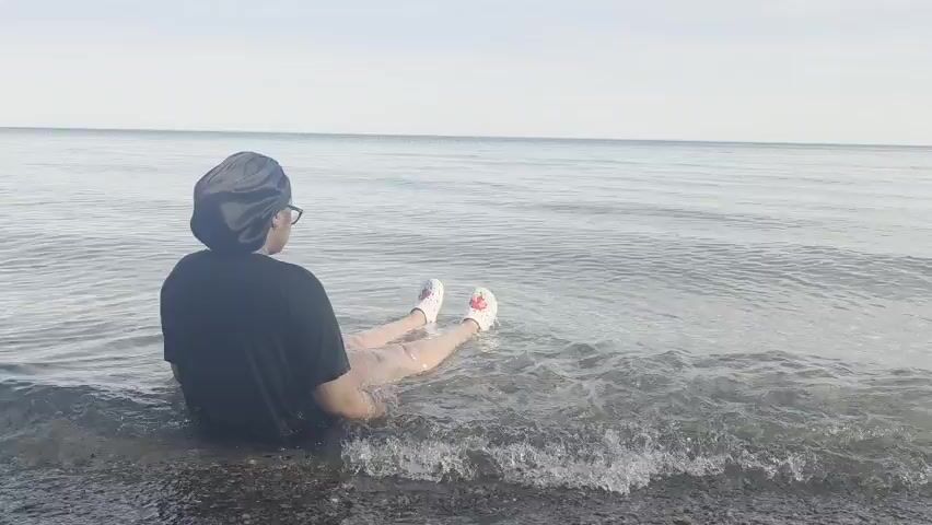 A person is shown from behind, sitting in the shallow water of a large body of water. They are wearing a black t-shirt, a dark head covering (possibly a bonnet), and glasses. Their lower legs and feet, which are clad in white shoes with red accents, are submerged in the water. Small waves gently lap around them. The person is facing away from the camera, looking out towards the horizon where the water meets a light, overcast sky. The scene is calm, suggesting a peaceful moment by the water, likely Lake Ontario given the location context of Ajax, Canada. The time of day appears to be daytime, under an overcast or hazy sky. There is no visible text.