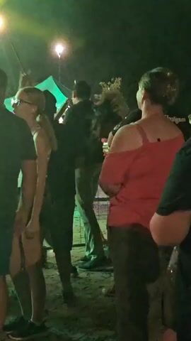 The video shows a crowd of people gathered outdoors at night. There are bright lights, including green and red, illuminating the scene. In the foreground, several people are visible from behind, facing away from the camera. One woman in a red off-the-shoulder top is prominent. To the left, a woman with blonde hair and glasses is also visible. In the middle ground, a man in a black shirt appears to be interacting with others, possibly dancing or moving. The background suggests a festival or event setting with tents and more people. The overall atmosphere appears energetic and social.