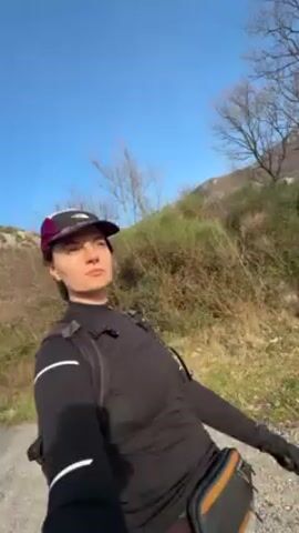 A woman is walking on a gravel path. She is wearing a black long-sleeved shirt, black pants, a small backpack, a fanny pack, and a baseball cap.  The sky is blue and the sun is out. There are trees and bushes on either side of the path. A hill can be seen in the background. The woman is looking around as she walks.