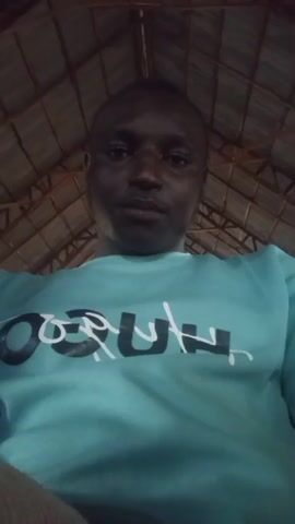 A man wearing a teal sweatshirt with the word "HUGO" printed on it smiles and looks around. The ceiling above him is made of thatched material supported by wooden beams.