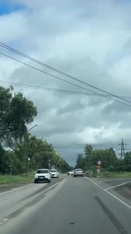 This media file captures a view from a vehicle driving on a road in Slantsy, Russia. The scene depicts a two-lane asphalt road flanked by lush green trees and vegetation on both sides. The sky is overcast with heavy, gray clouds interspersed with patches of blue, suggesting a mixed weather day, possibly late afternoon given the diffused light.

Several cars are visible on the road. In the foreground, a white SUV is driving ahead, followed by another white car. Further down the road, a silver sedan is also in motion. On the right side of the road, near a cluster of trees, a traffic sign with the number "30t" and another with "20" are clearly visible, indicating weight and speed limits respectively. These signs, along with the general appearance of the road and surrounding foliage, are consistent with a rural or semi-rural setting in Russia.

The road itself shows signs of wear, with some unevenness and patching visible, particularly in the foreground. Power lines are strung across the sky, supported by poles, adding to the utilitarian aspect of the landscape. The overall atmosphere is that of everyday travel through a somewhat undeveloped, green area. There are no people clearly visible as subjects within the frame, focusing instead on the road and vehicular activity. The time of day appears to be daytime, but the cloudy conditions create a somewhat subdued lighting.