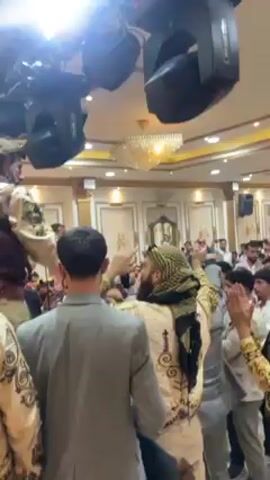 The video shows a large gathering of people inside a decorated hall. The focus is on a group of men wearing traditional Middle Eastern clothing, with black-and-white keffiyehs and ornate vests. They are dancing and clapping their hands, seemingly celebrating. The hall has ornate paneling, a chandelier, and stage lights. Other people in the crowd are clapping and cheering along.