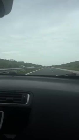 The video shows a car driving on a highway. The camera is positioned inside the car, looking out through the windshield. There are other vehicles on the road. The sky is overcast. There is vegetation on either side of the highway, with a guardrail visible on the left side. The car's dashboard is visible in the foreground.