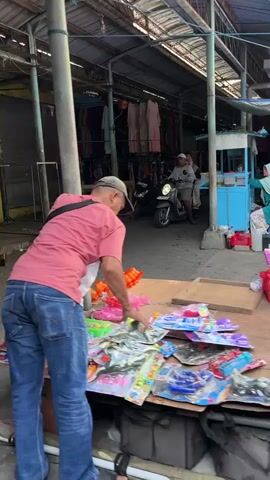 A man wearing a pink shirt and blue jeans is seen at an outdoor market stall. He is reaching down to pick up items from a table covered in toys. The toys are brightly colored and packaged. In the background, a person on a scooter is visible, and there are more stalls with goods displayed. The scene appears to be during the daytime.