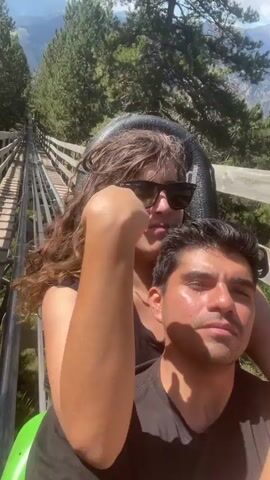 This video shows a close-up, selfie-style shot of a man and a woman in a roller coaster cart. The man is in the foreground, with his face tilted up slightly towards the camera. He is wearing a black t-shirt. The woman is behind him, with her arms around his neck. She is wearing a black t-shirt and sunglasses. Her hair is long and curly. She is smiling and looking at the camera. The background shows a wooden track for the roller coaster, surrounded by pine trees and mountains. The sky is blue with some clouds. The video appears to be taken during the daytime.