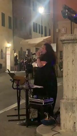 The video shows a street scene at night with a live musical performance. A woman is singing into a microphone on a stage with musical equipment. In the background, people are seated at tables outside a restaurant, and a couple is dancing in the street. The scene is illuminated by streetlights and colorful stage lighting.