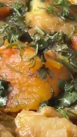 The video shows a close-up shot of a stew-like dish, filled with soft, orange-colored vegetables and green herbs, within a decorative metal serving dish.  The camera then pans out, revealing the dish to be part of a makeshift meal arrangement on a spread on the floor. Around the dish are other bowls, water bottles, bread, a spoon, and a small container of red sauce. In the background, there is a shelf with various items, a chair, and a glimpse of a door.  The lower portion of the frame shows the legs of a person wearing dark clothing.