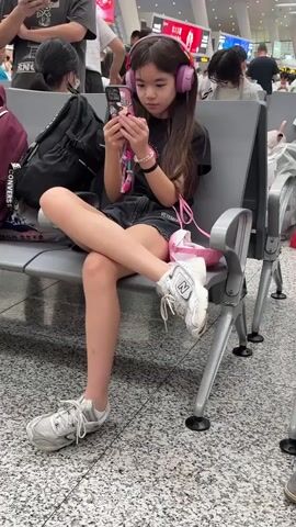 A young girl with long, dark hair is sitting on a bench in what appears to be a train station or airport waiting area. She is wearing large pink headphones and is looking down at her phone, which she holds in both hands. Her left leg is crossed over her right leg, and she is wearing a black t-shirt, denim shorts, and white sneakers. A pink bag is on the bench next to her. Other people are visible in the background, sitting on benches and walking around. The environment appears to be indoors, with a tiled floor and gray seating.
