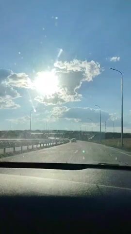 A first-person perspective video captures a journey along a highway under a bright, partly cloudy sky. The sun is shining intensely, creating glare on the windshield. The road ahead is paved and appears to be a multi-lane highway with guardrails on the left. Several streetlights are visible along the sides of the road, and a bridge or overpass is prominent in the distance. A few cars can be seen on the road ahead. The dashboard of the vehicle is partially visible in the foreground. The overall impression is of a bright, sunny day and a smooth drive.