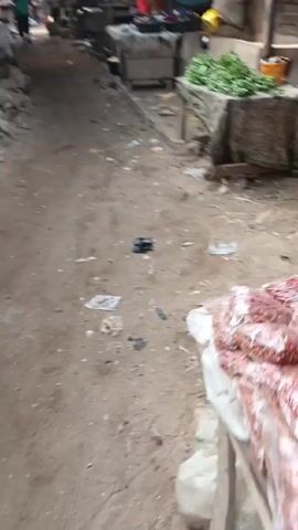 The video shows a narrow alleyway in an outdoor market. The ground is dirt and scattered with trash. There are stalls on either side, some with merchandise visible, including leafy green vegetables and plastic goods. Several people can be seen walking and standing in the alleyway. One woman is wearing a red dress and pink shoes. The market appears to be under a corrugated metal roof.
