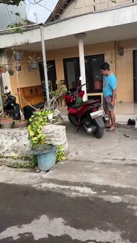A person in a blue t-shirt is seen sitting on a red motorcycle parked outside a building. The motorcycle is positioned in front of some plants and a concrete structure. The building appears to be a house with a tiled roof and yellow walls. The ground is paved with concrete and asphalt.