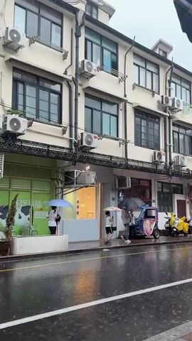 A still shot shows a street scene on a rainy day. The pavement is wet and reflects the light. In the background, a row of multi-story buildings with cream-colored facades and dark windows is visible. Each window has an air conditioning unit installed below it. There are several storefronts at street level. One on the left has a green facade with a cartoon cow image and text. Another in the center is a lighter color with a glass door. A third on the right has a red accent around its doorway. People are walking on the sidewalk, most of whom are holding umbrellas. A person on a scooter with a purple poncho rides across the frame from left to right. A three-wheeled vehicle with colorful decorations is parked on the right side of the street, with a yellow scooter parked behind it. The overall atmosphere is wet and overcast.