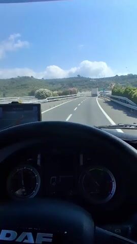 The video shows a first-person perspective from the driver's seat of a truck. The driver is on a highway with a clear blue sky and some clouds overhead. The road is asphalt with white lane markings and a metal guardrail on the right side. There are hills and vegetation on either side of the highway. The truck's dashboard and steering wheel are visible in the foreground, with a GPS device mounted on the dashboard. Other trucks are visible in the distance on the highway. The driver's hands are on the steering wheel.